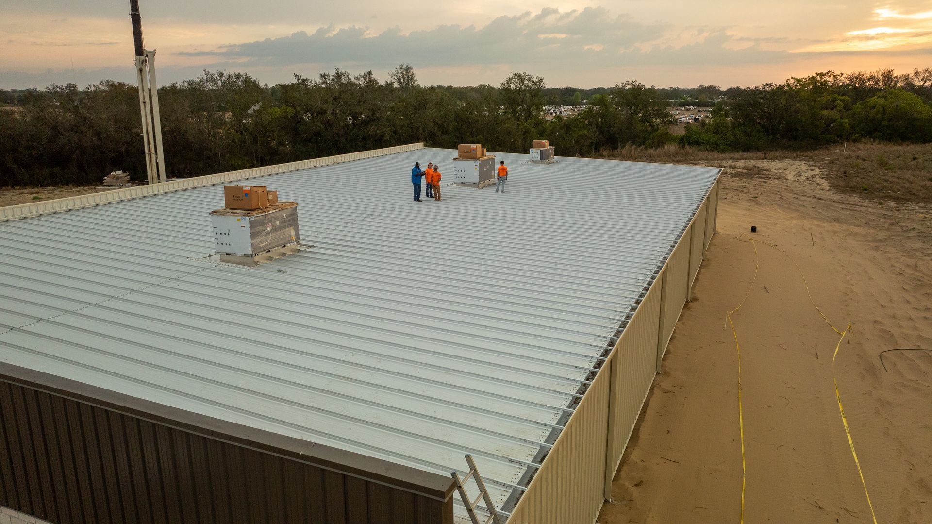 Rooftop of a corrugated metal building with several vents, surrounded by sandy ground and trees at sunset