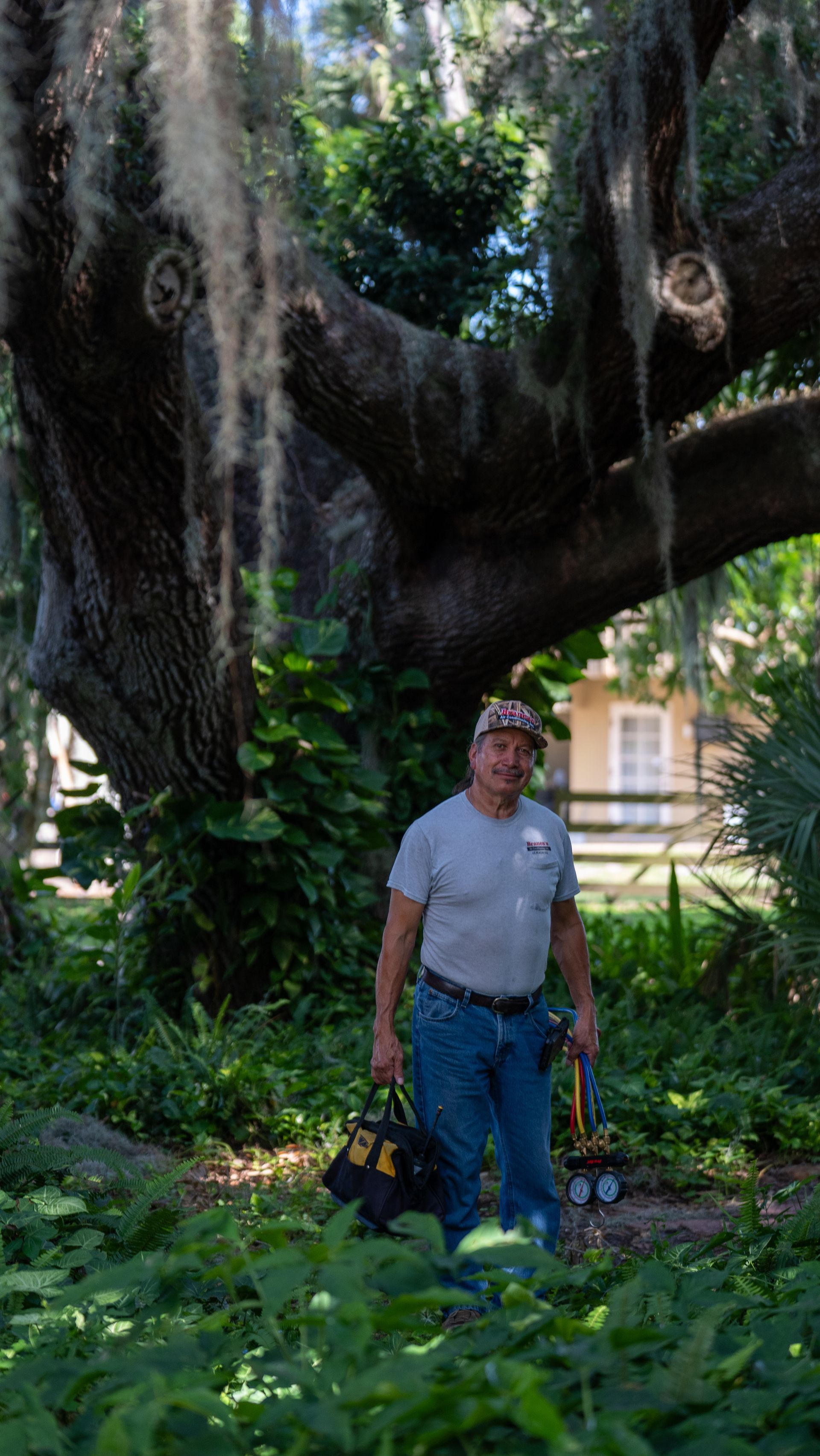 Man standing under a large tree with hanging moss in a sunny garden, holding tools beside a bag.
