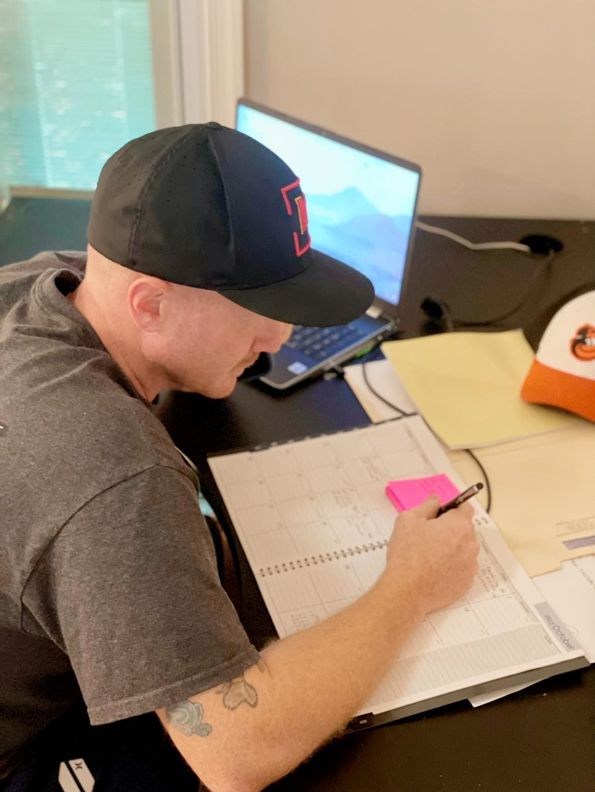 A man wearing a hat is sitting at a desk writing in a notebook