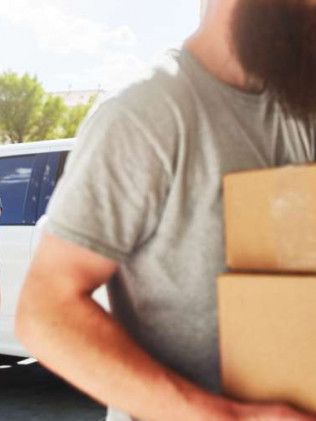 Man carrying two cardboard boxes, standing next to a white vehicle, sunny outdoors.