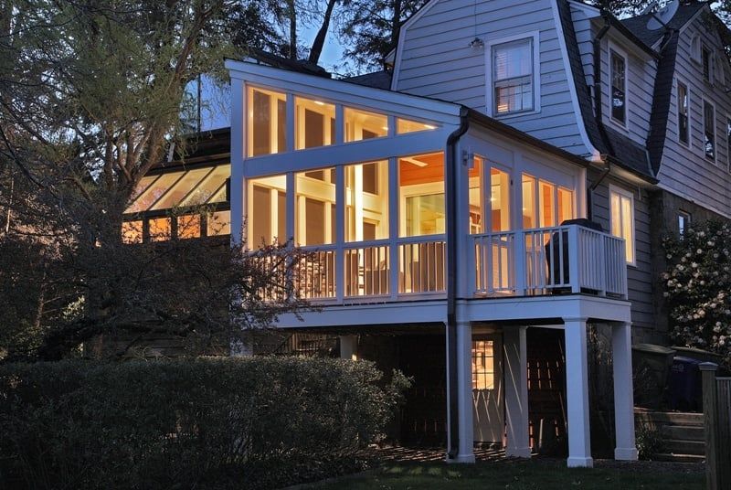 Lit sunroom extension on a two-story house, with white railings and pillars; exterior lit at dusk.