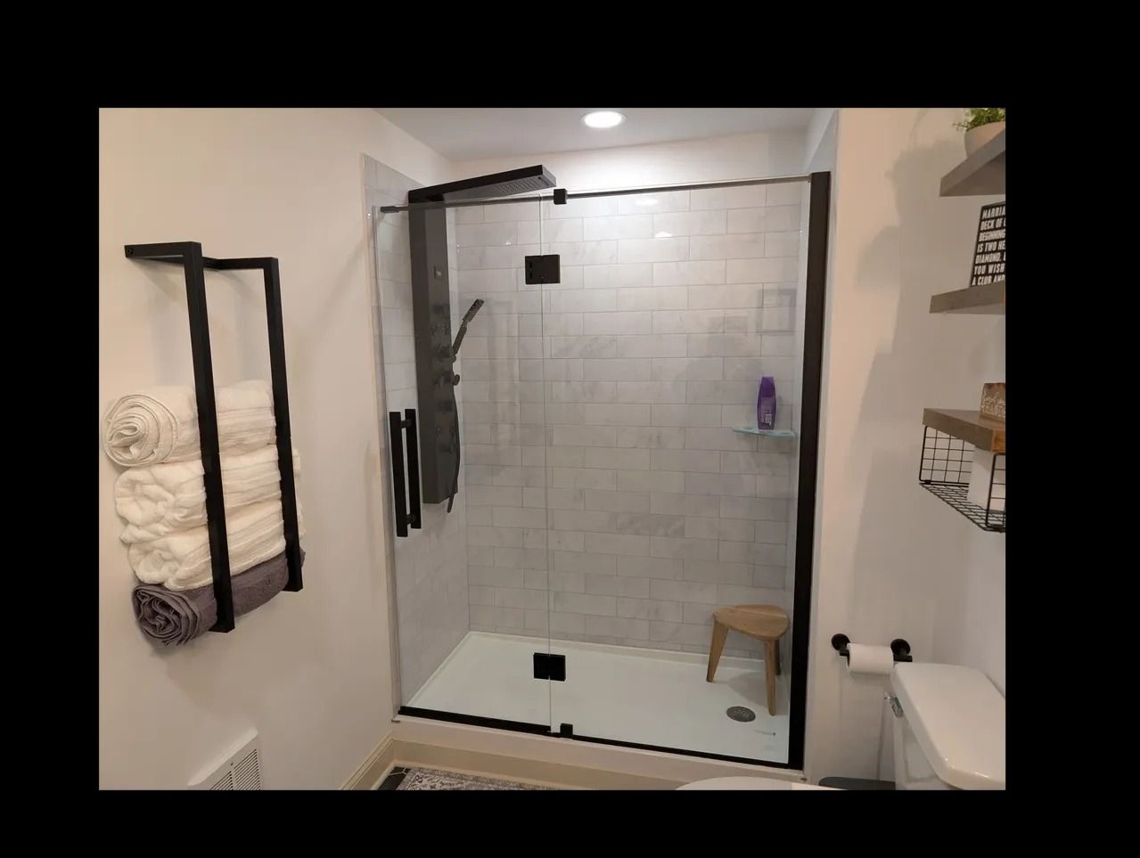 Modern bathroom with a glass shower, white tile, black trim, and a towel rack with folded towels.