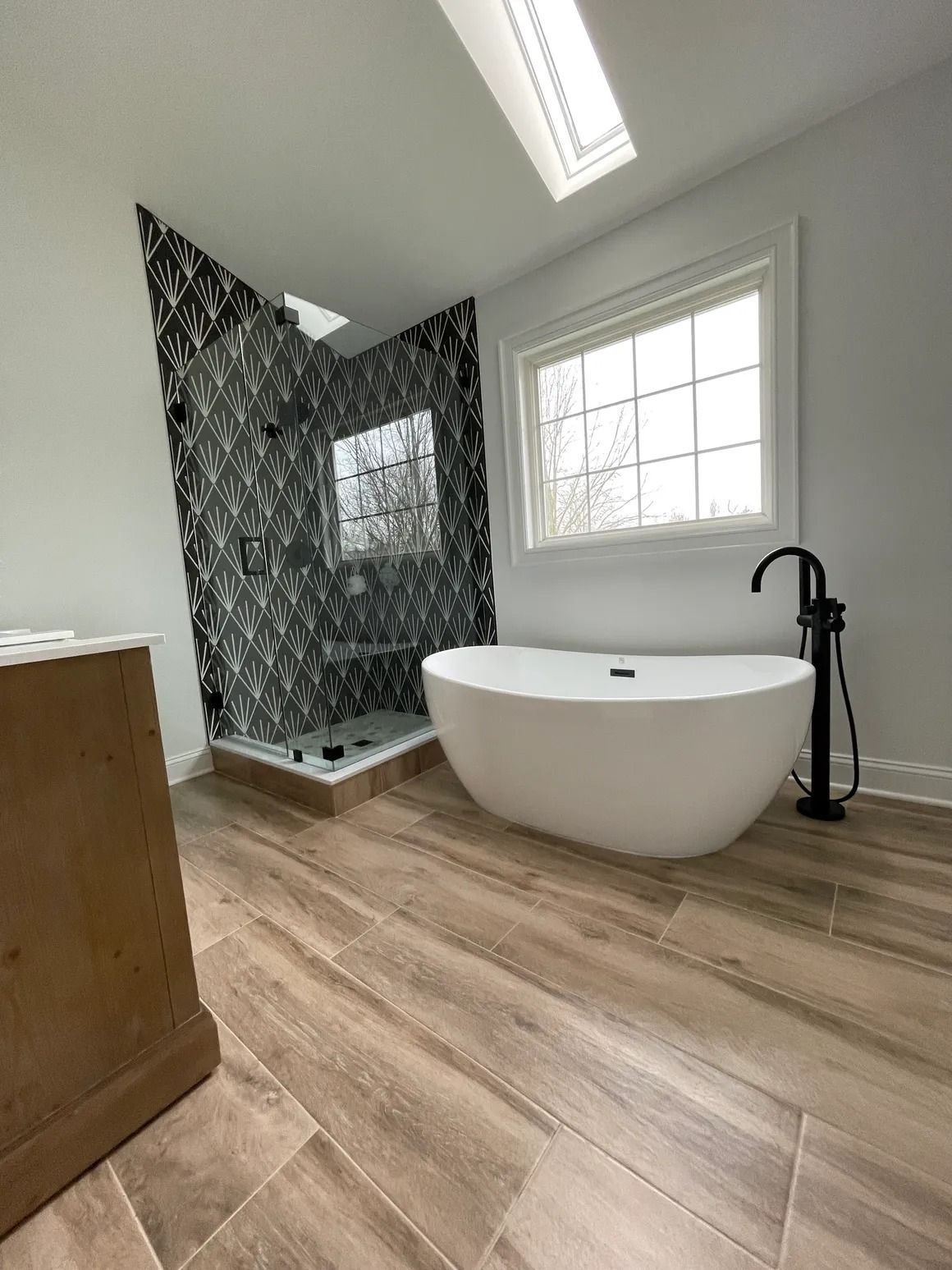 Bathroom with patterned shower, freestanding tub, window, and wood-look tile floor.