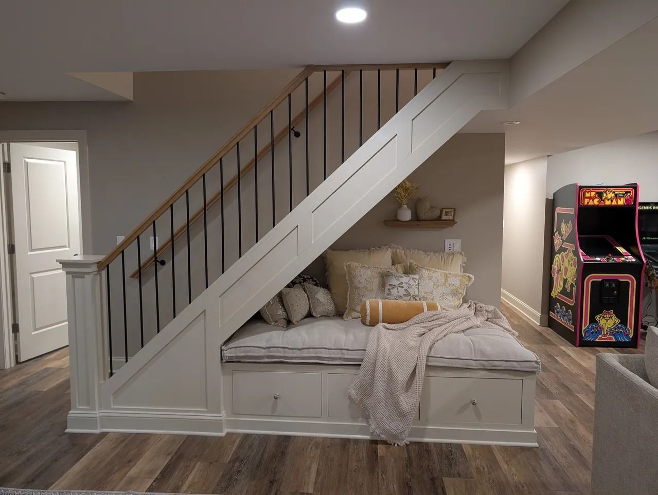 Cozy basement seating area under stairs with built-in storage, cushions, pillows, and a blanket. An arcade game is visible.