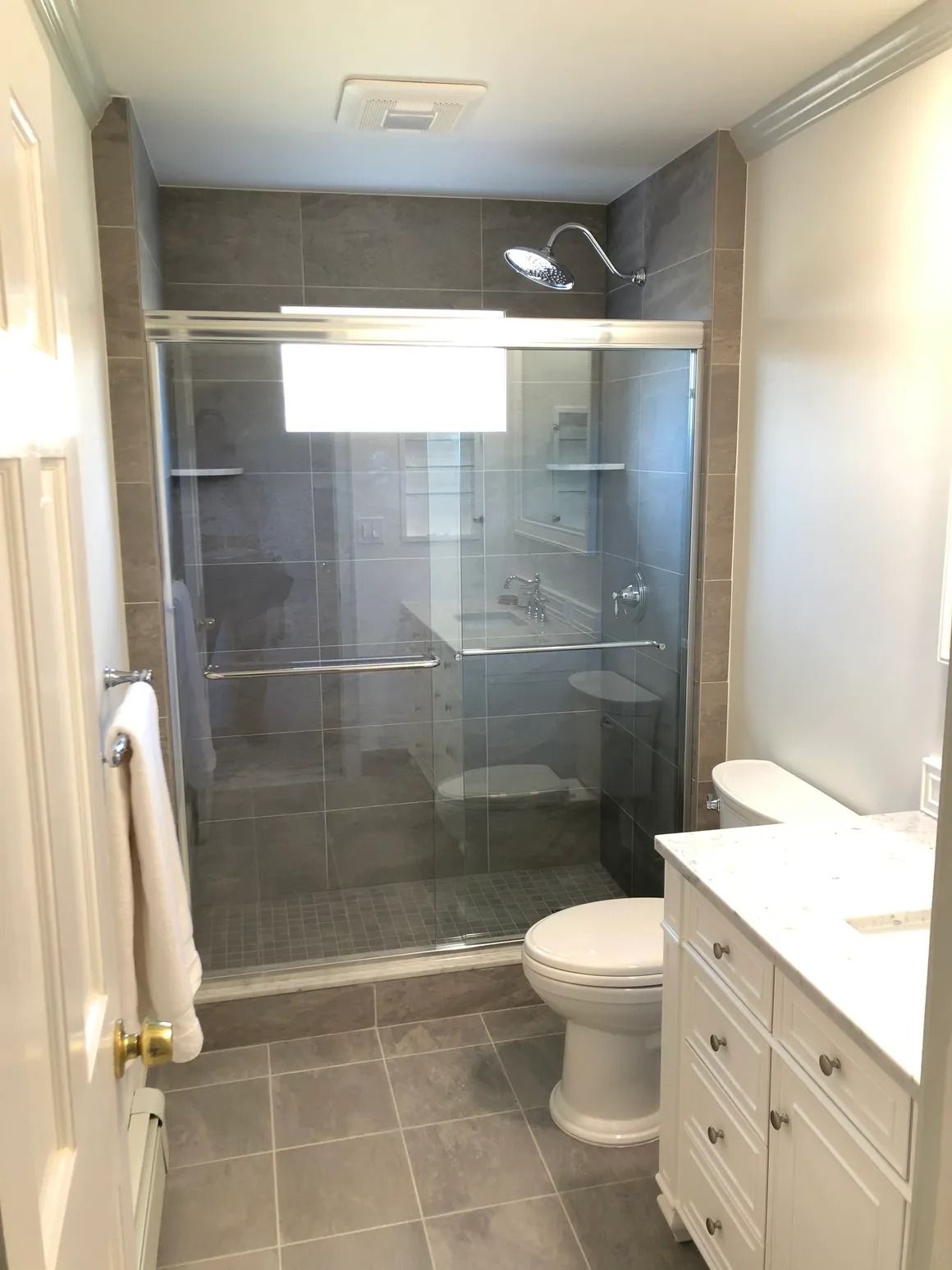 Bathroom with glass shower, gray tiled walls/floor, white vanity/toilet, and a towel on a door.