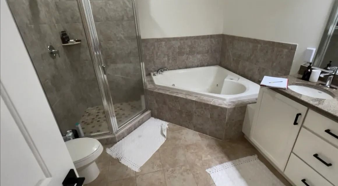 Bathroom with shower, corner tub, toilet, and vanity. Beige tile walls and floor, white cabinets.