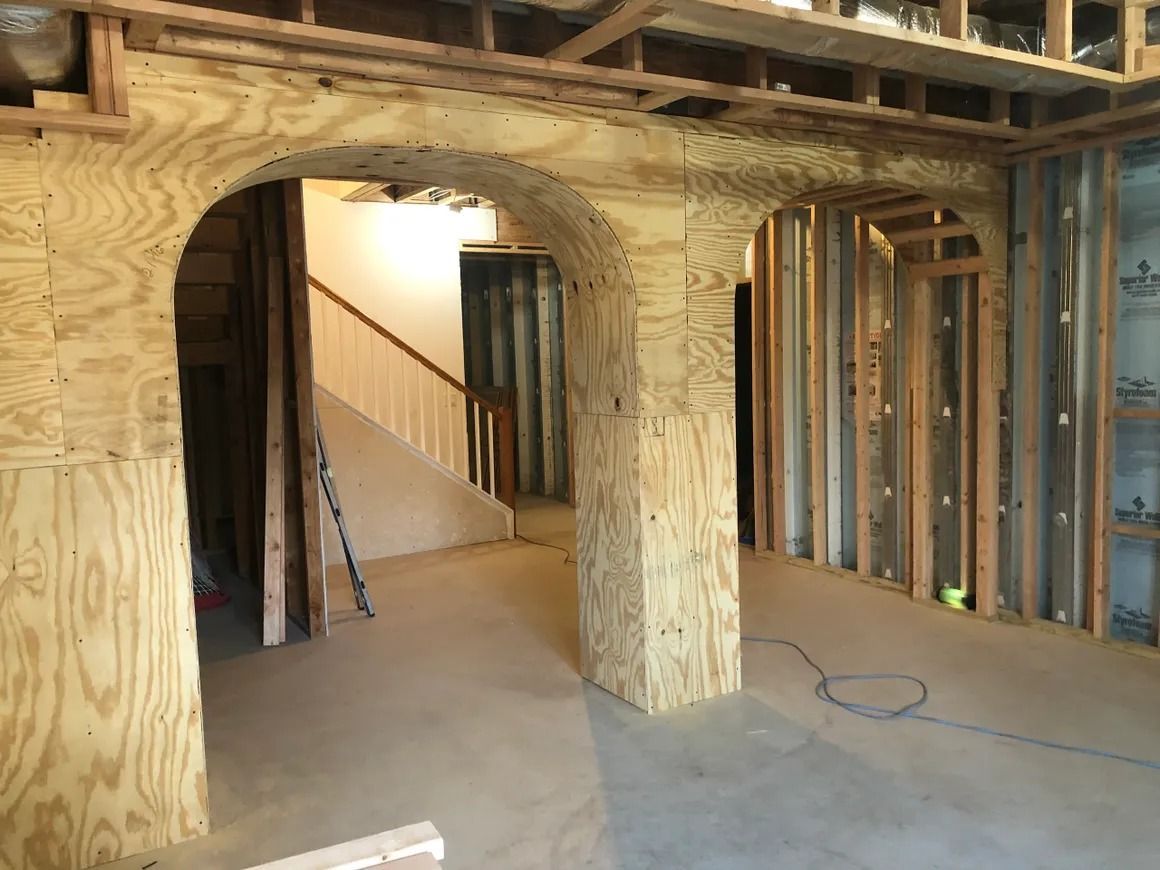 Interior of a room under construction with plywood archways, exposed studs, and a staircase in the background.