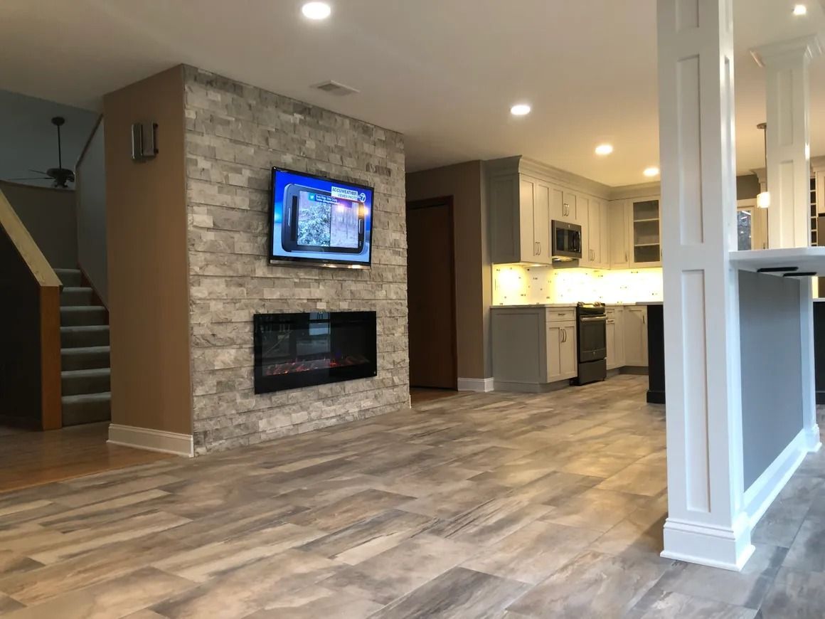 Basement living area with a fireplace, TV, and kitchen. Light gray cabinets, hardwood-look flooring, and a staircase.