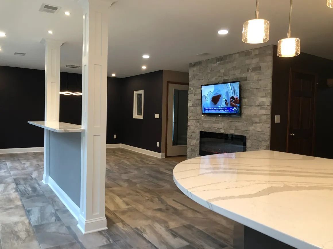 Modern finished basement with bar, fireplace, and TV. Grey stone accent wall, white and grey countertops, dark blue walls.