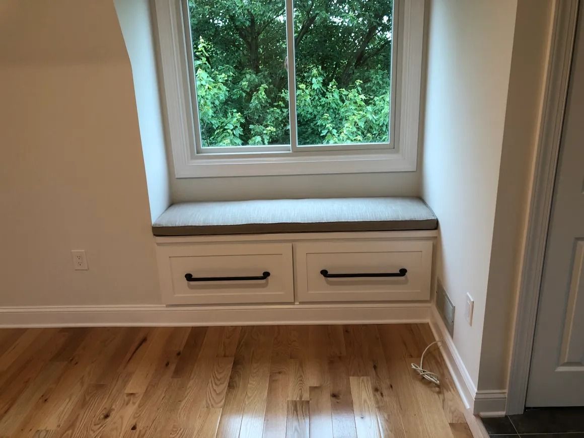 Window seat with drawers, gray cushion, white frame, hardwood floor, window with green foliage outside.