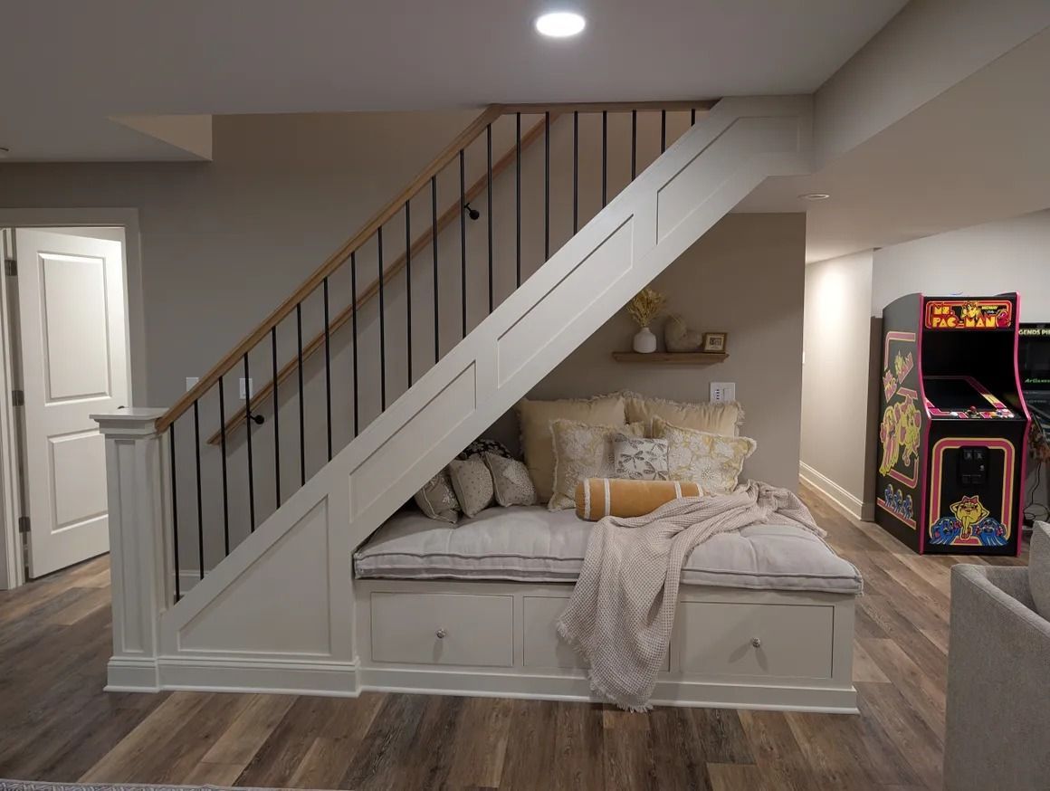 Cozy reading nook built under stairs. Features built-in storage, cushioned seat, pillows, and a blanket. Arcade game visible.