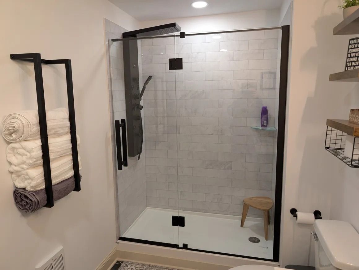 Bathroom with a white-tiled shower, black trim, glass door, towels on a wall rack, and a small stool.