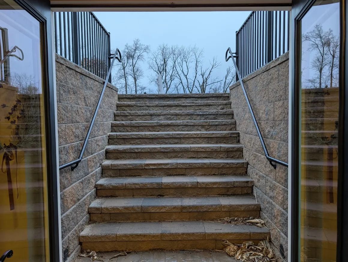 Stone staircase leading up, viewed through open doors. Black railings, trees in background, cloudy sky.