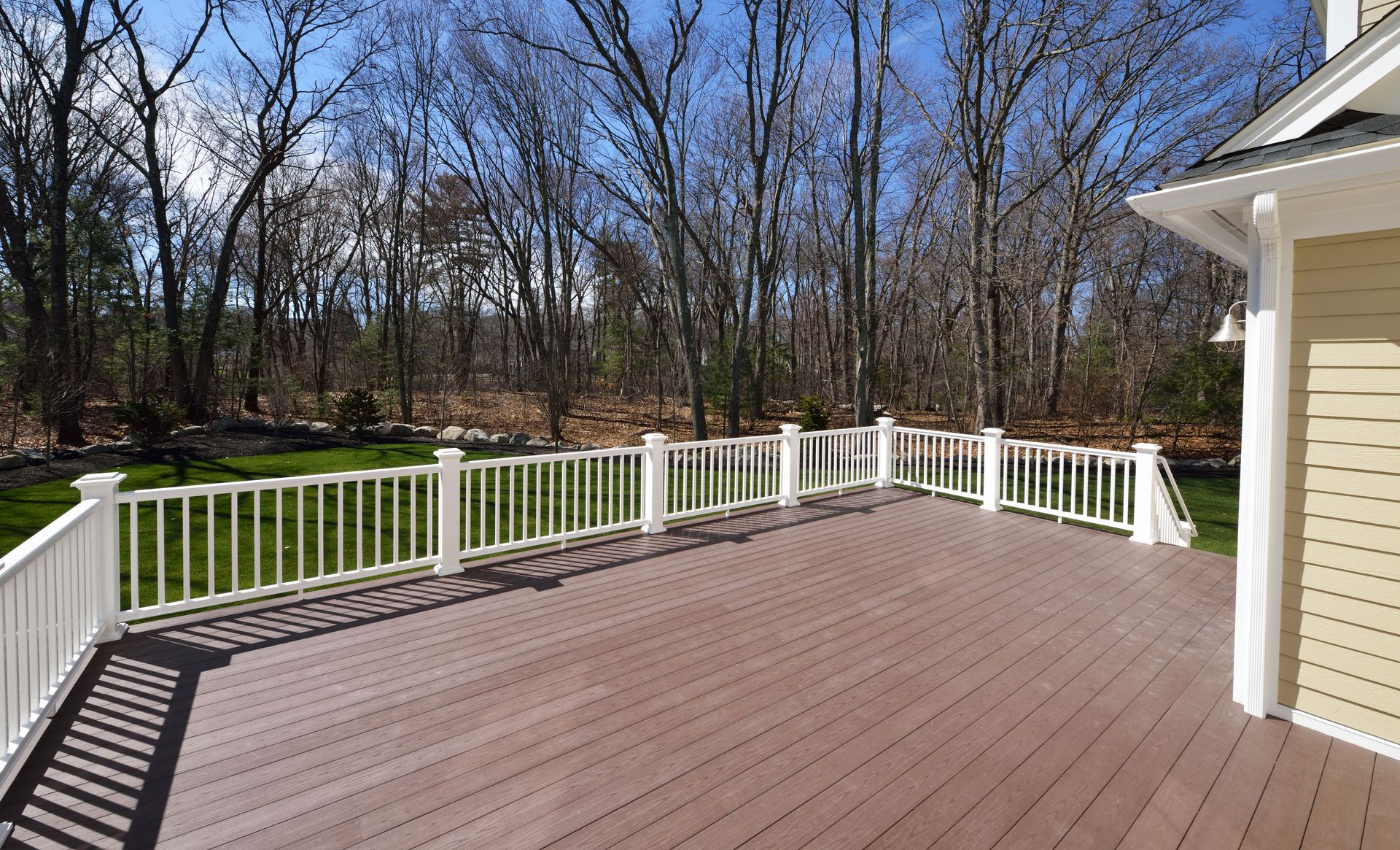Wooden deck with white railing, overlooking a wooded area. The house has tan siding.
