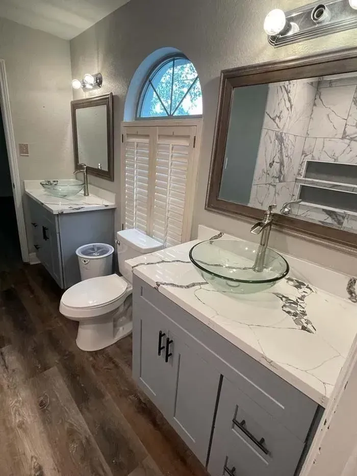 Modern bathroom with double sinks, marble countertops, large mirror, toilet, and arched window with shutters