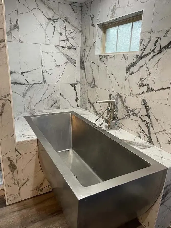 Stainless steel bathtub in a marble-tiled bathroom with a small frosted window.