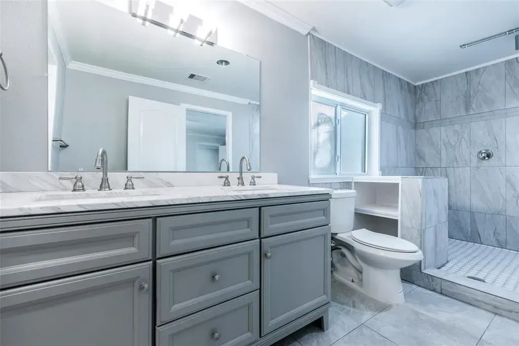 Bright modern bathroom with gray vanity, large mirror, toilet, shower, and marble tile walls and floor