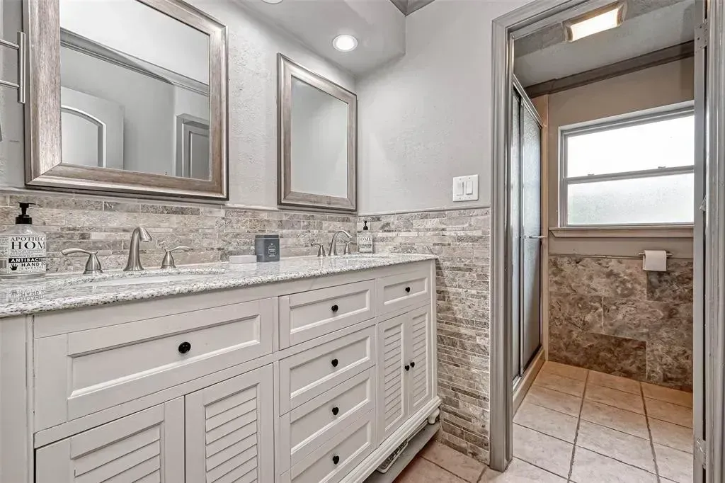 Bright bathroom with double vanity, large mirror, tiled walls, and open doorway to a shower area