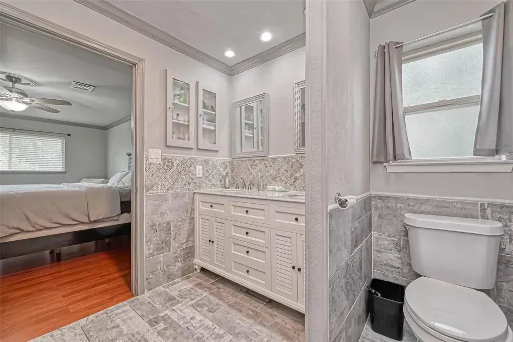 Bright bathroom with white vanity, toilet, gray tile, and a mirrored wall beside a bedroom.