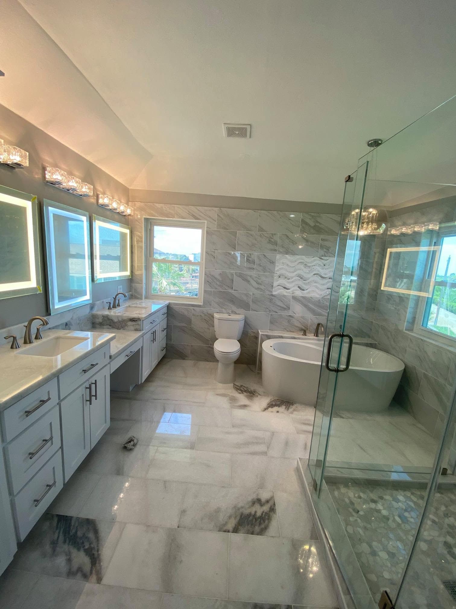 Bright bathroom with double vanity, large windows, glass shower, freestanding tub, and white marble floor.