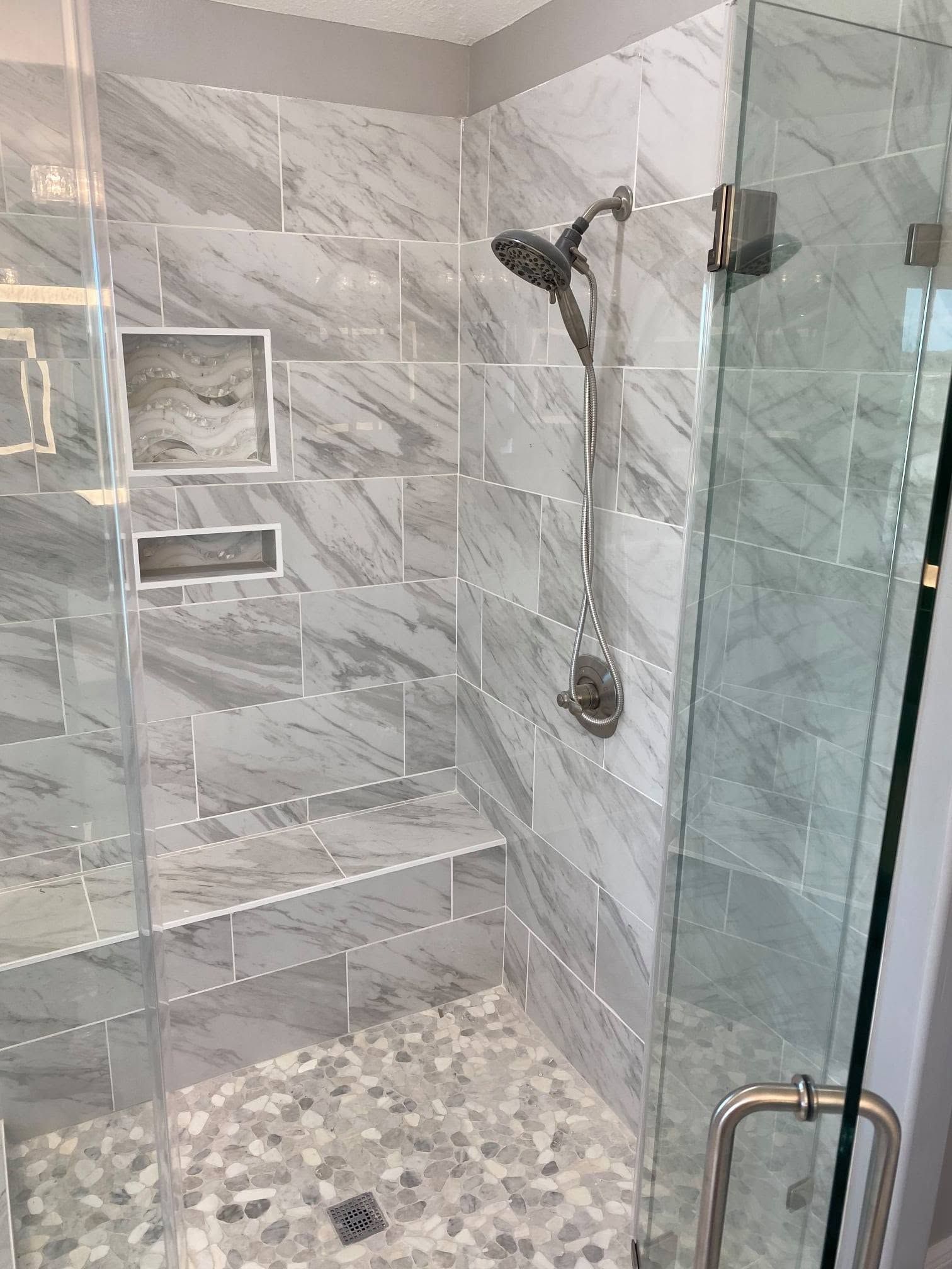 Modern tiled walk-in shower with glass door, rain showerhead, niche, and pebble floor.