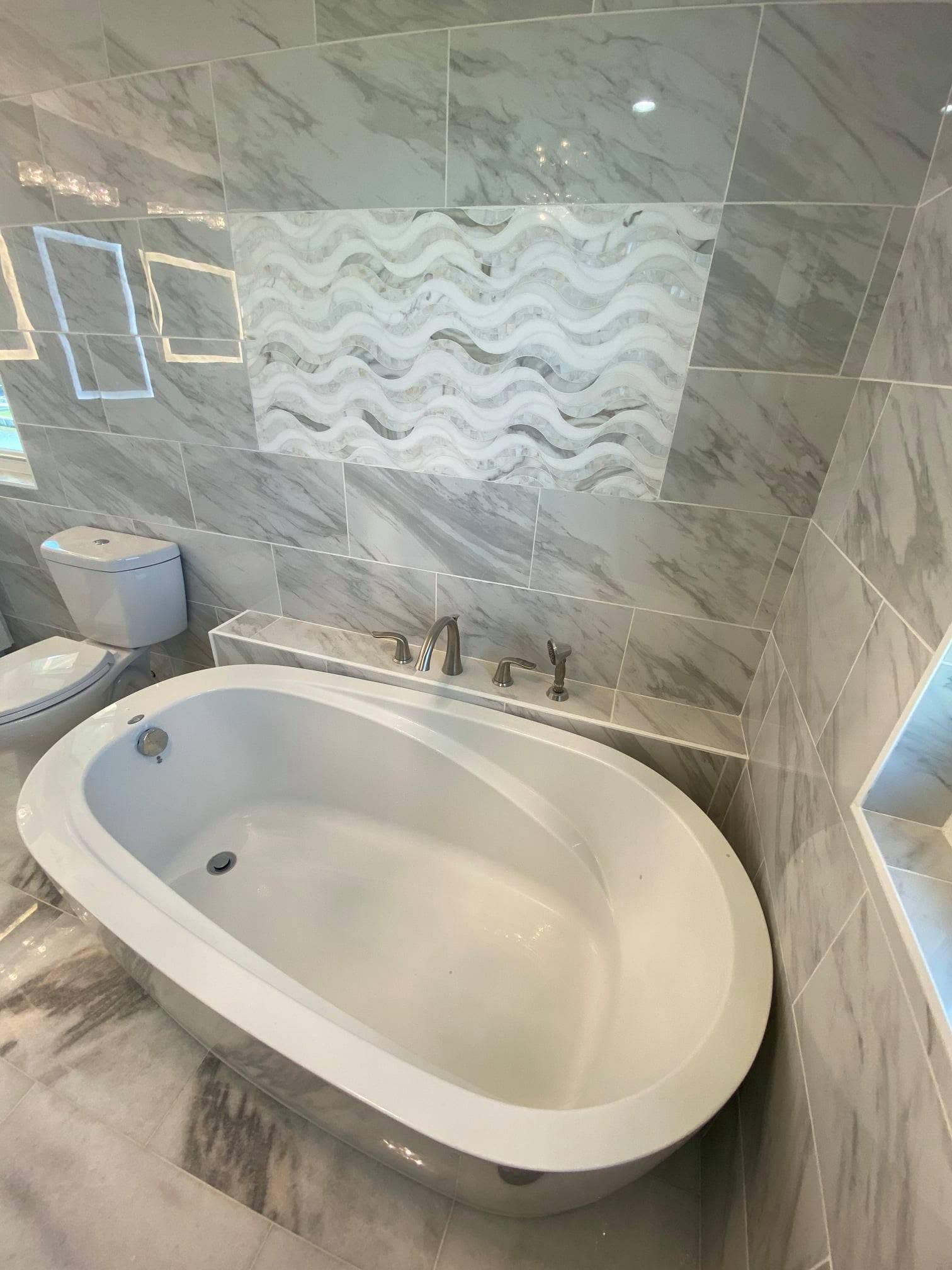 White oval bathtub in a marble-tiled bathroom with a decorative backsplash and chrome fixtures