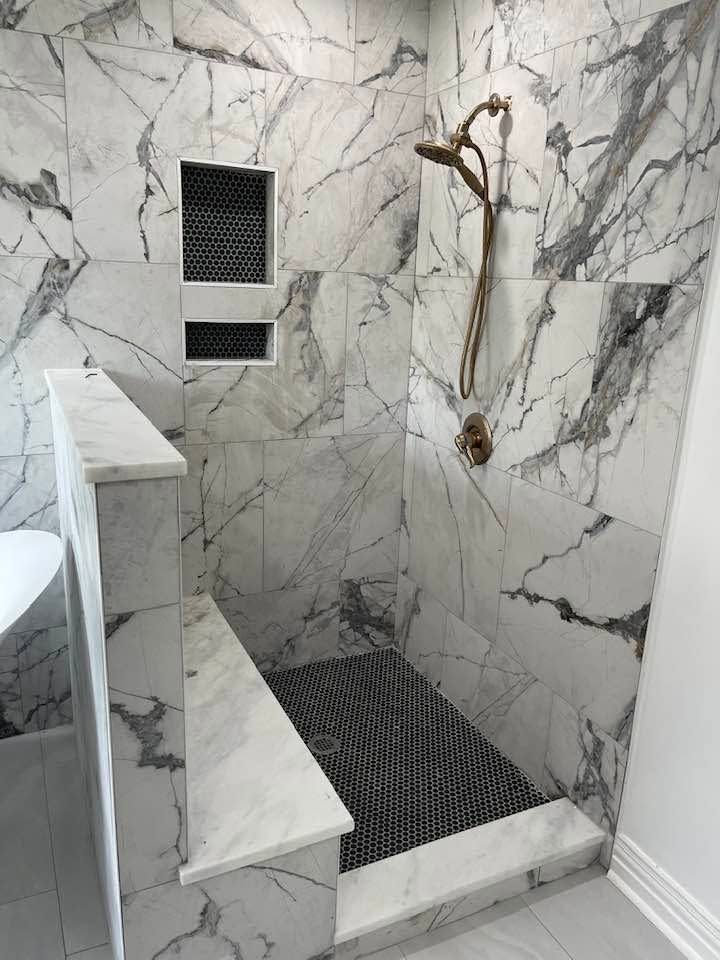 Small tiled shower with marble-look walls, built-in bench, floor drain, and handheld showerhead