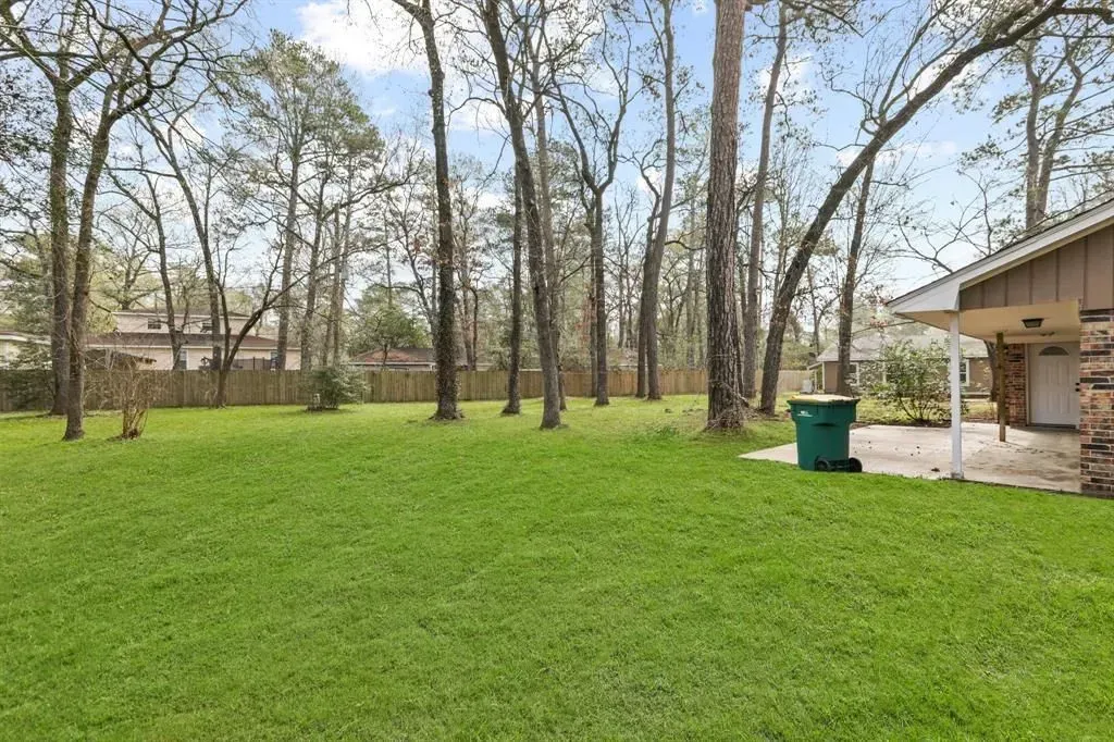 Grassy backyard with tall trees, a house patio on the right, and a green trash bin near the wall.