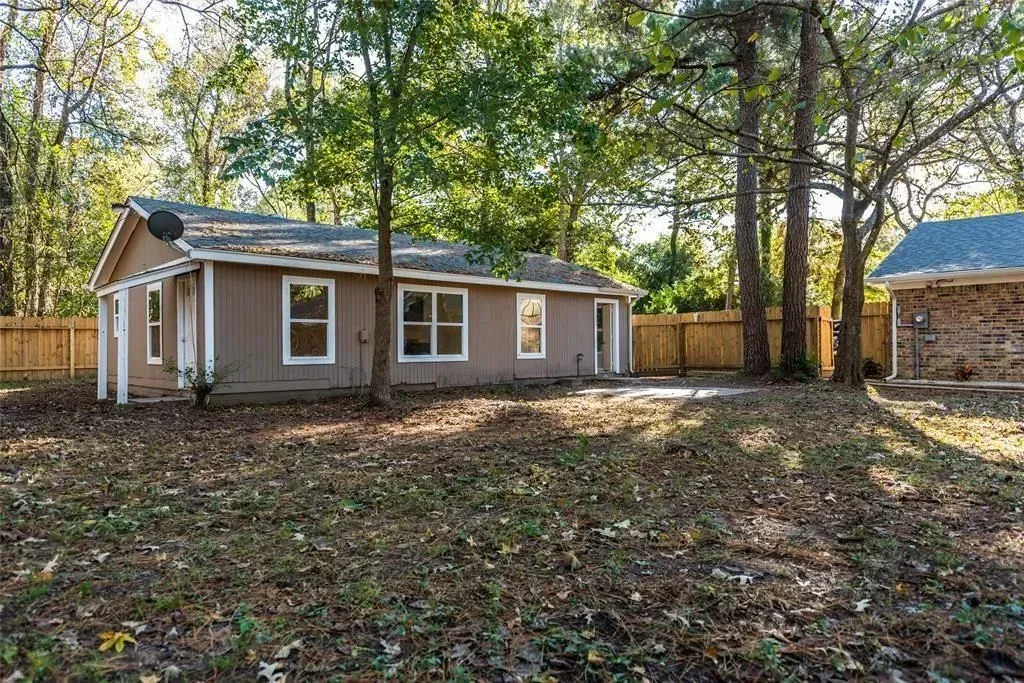 Small beige house with white trim in a wooded backyard, fenced and covered in fallen leaves.