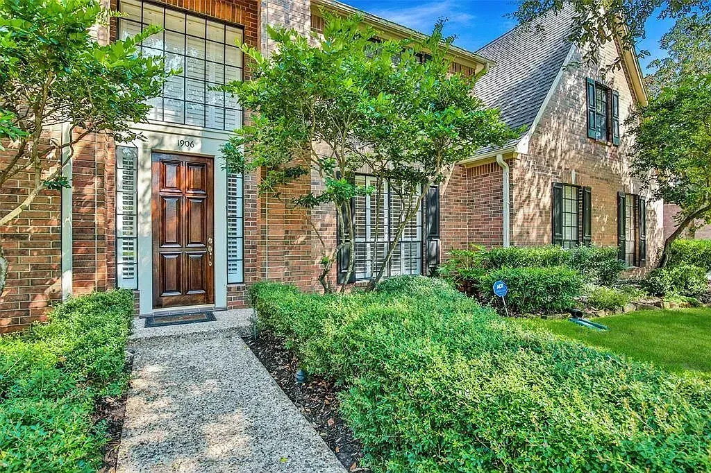Brick house entrance with a wooden door, stone walkway, and lush green landscaping