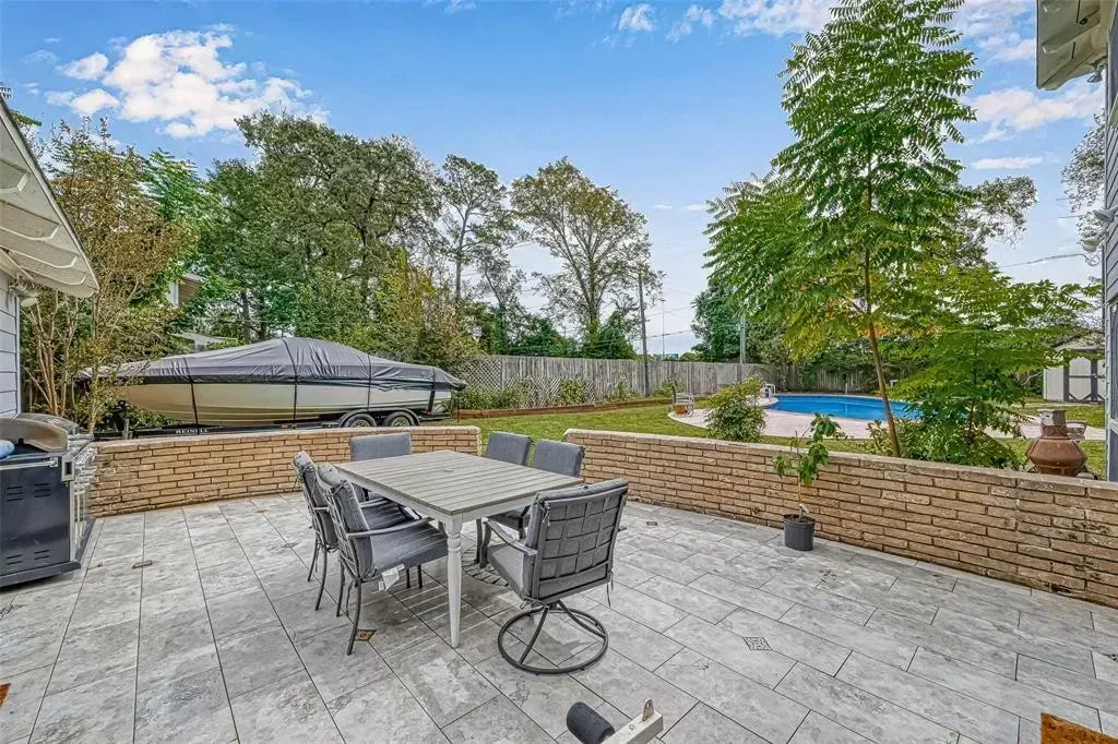 Patio with dining table and chairs, stone seating wall, trees, and a pool under a blue sky