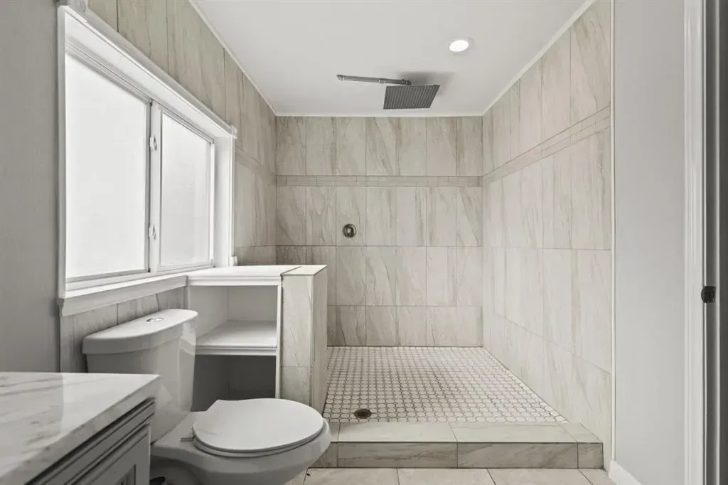 Small bathroom with toilet, window, and tiled walk-in shower in light beige tones