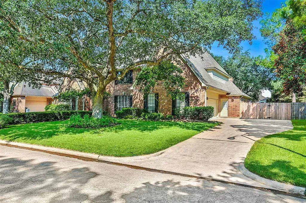 Tree-shaded house with a green lawn, curved driveway, and blue sky in a suburban neighborhood
