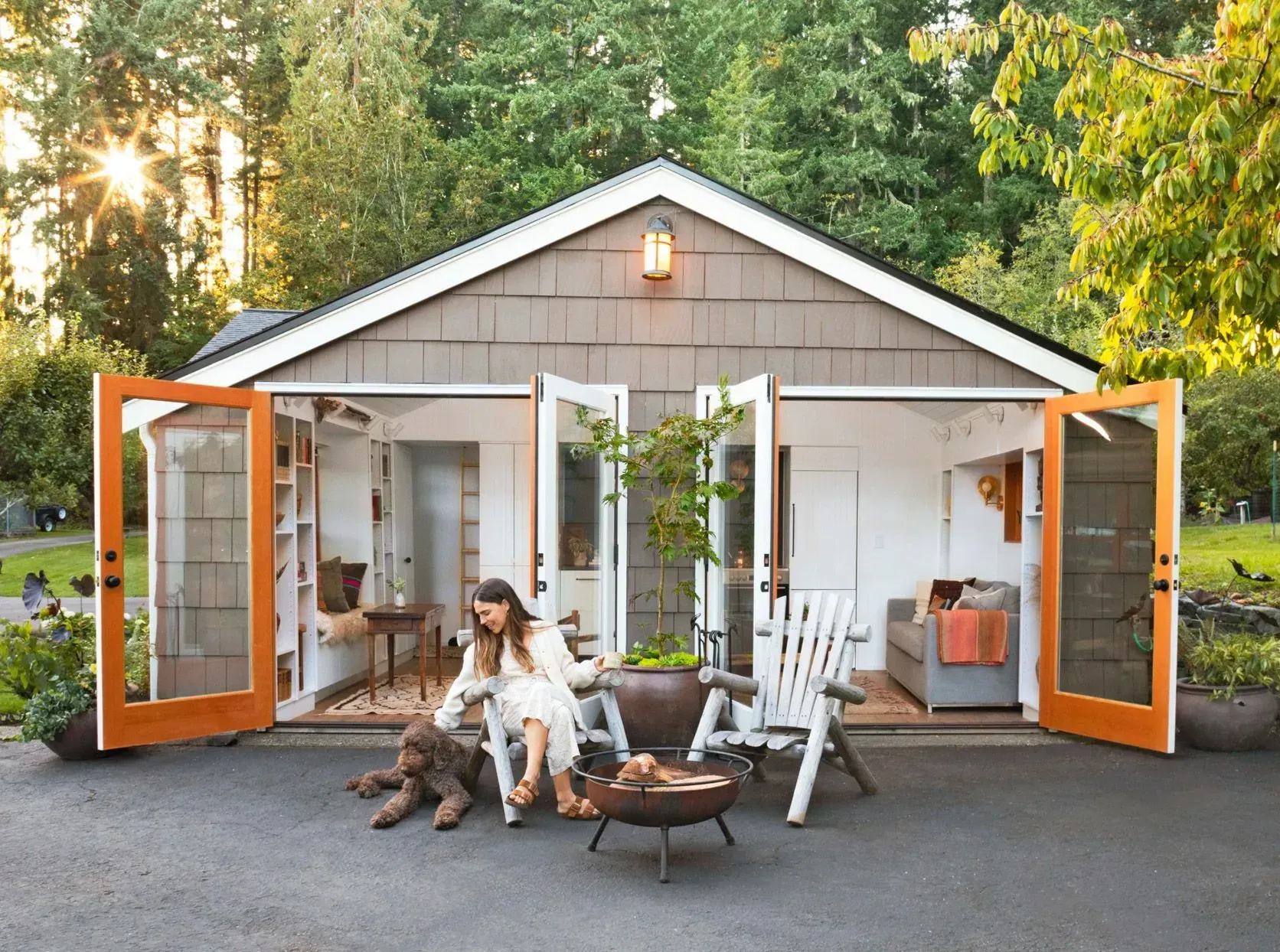 Open garage with orange doors, woman seated by dog, outdoor chairs, and a small shed-like workspace