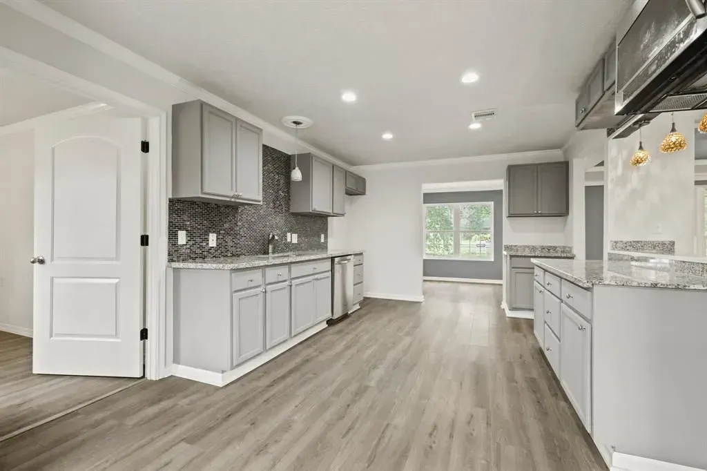 Bright modern kitchen with white cabinets, gray countertops, and wood-look flooring, viewed toward a windowed dining area