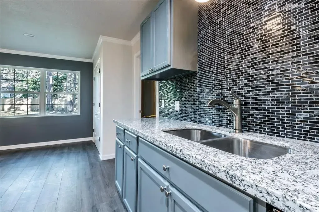 Modern kitchen with gray cabinets, black tile backsplash, and windowed dining area beyond