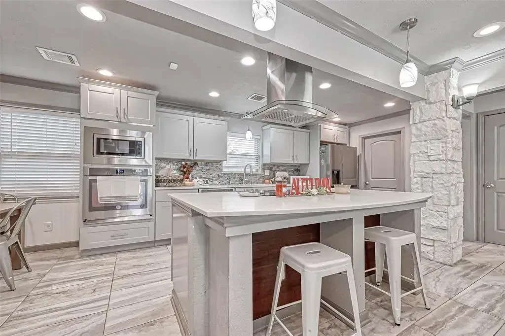 Bright modern kitchen with marble island, white stools, stainless appliances, and gray cabinetry