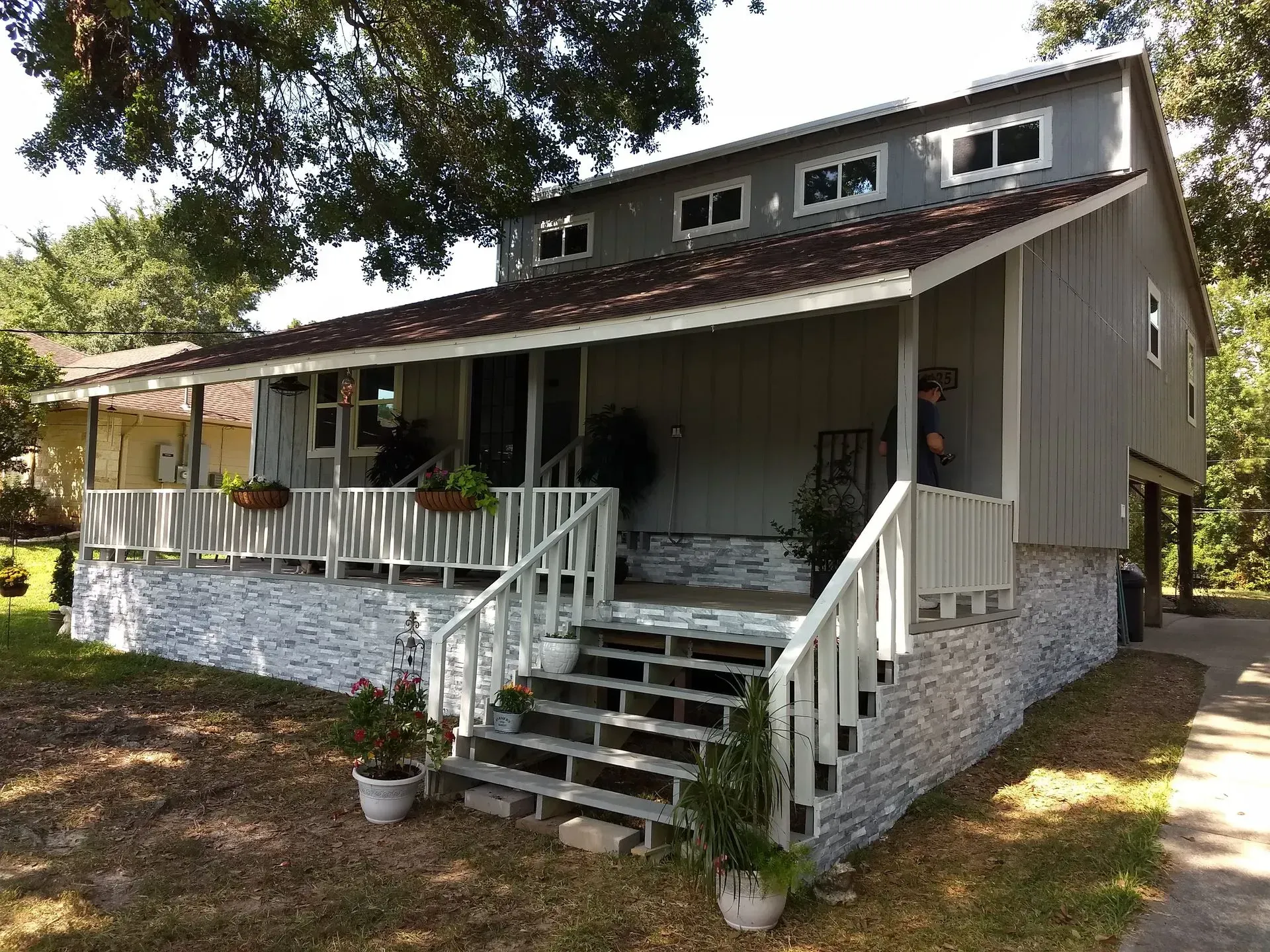 Two-story gray house with front porch, stairs, and stone foundation under trees