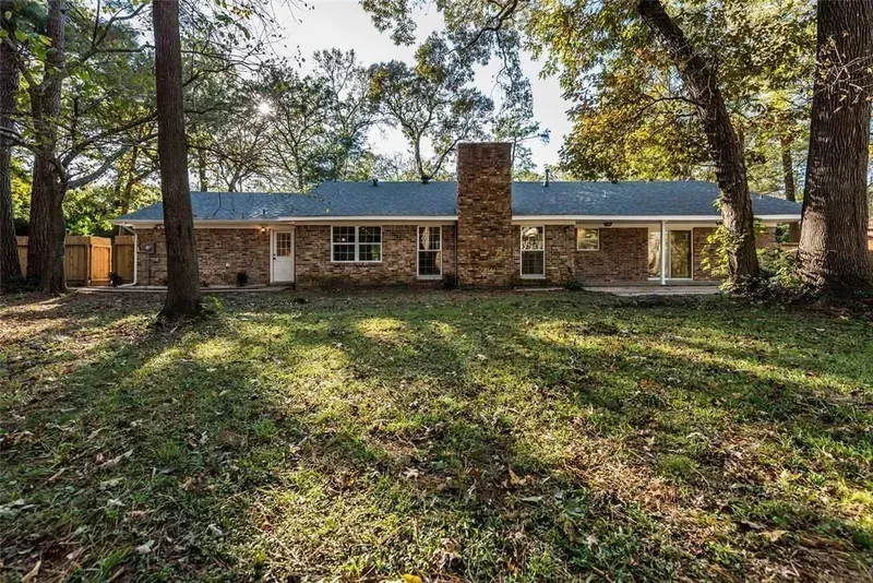 Single-story brick house with a central chimney, framed by large trees and a grassy front yard.