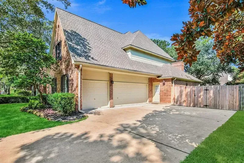Suburban brick house with two-car garage, driveway, green lawn, and tall trees on a sunny day
