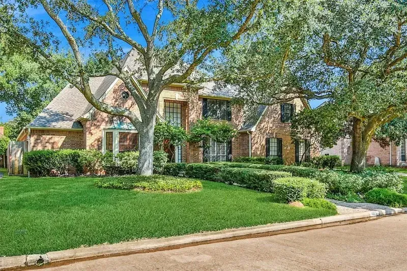 Suburban brick house with manicured lawn, trees, and a curved driveway under a blue sky