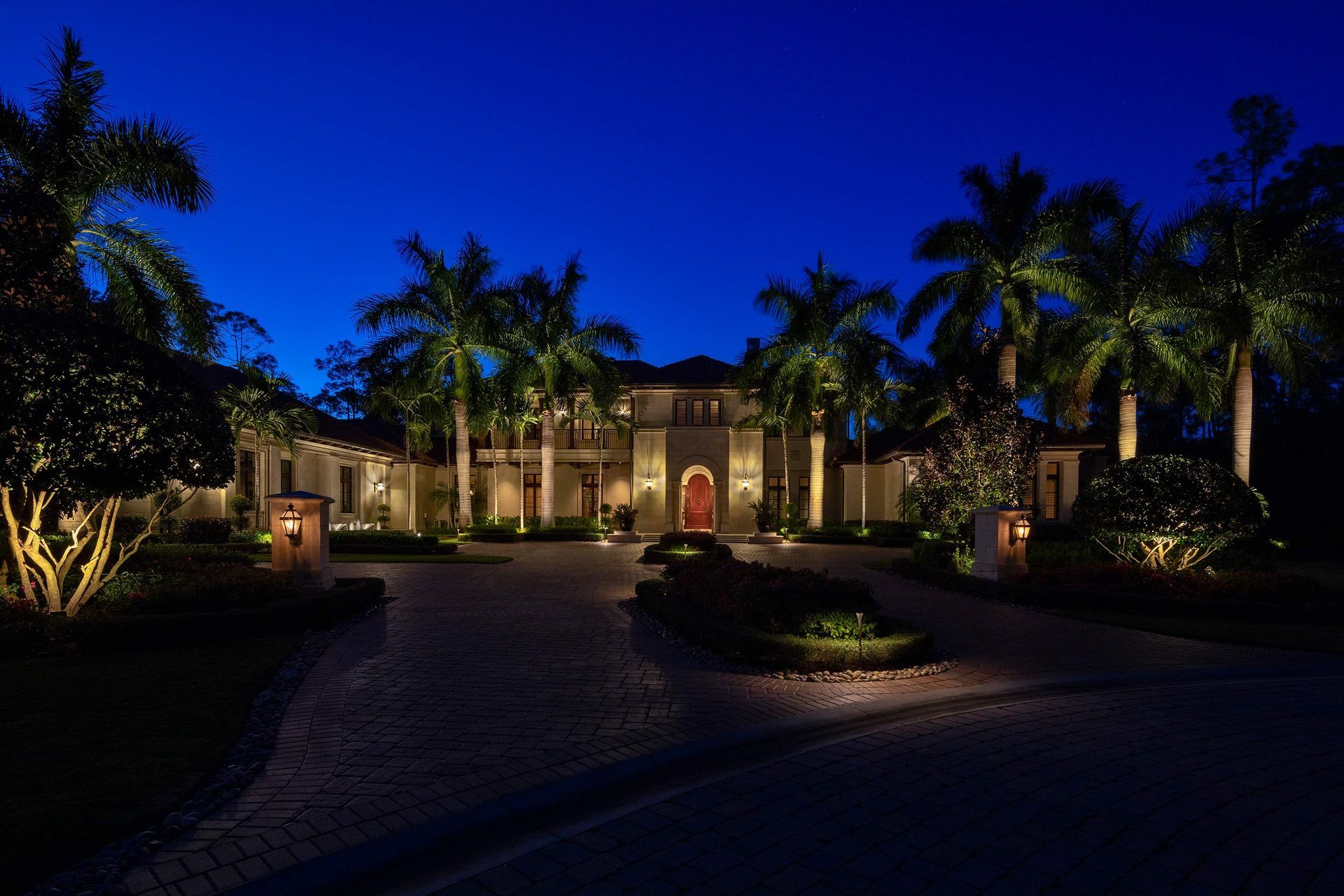 Night view of a luxurious house with palm trees, driveway and garden lights under a dark blue sky.