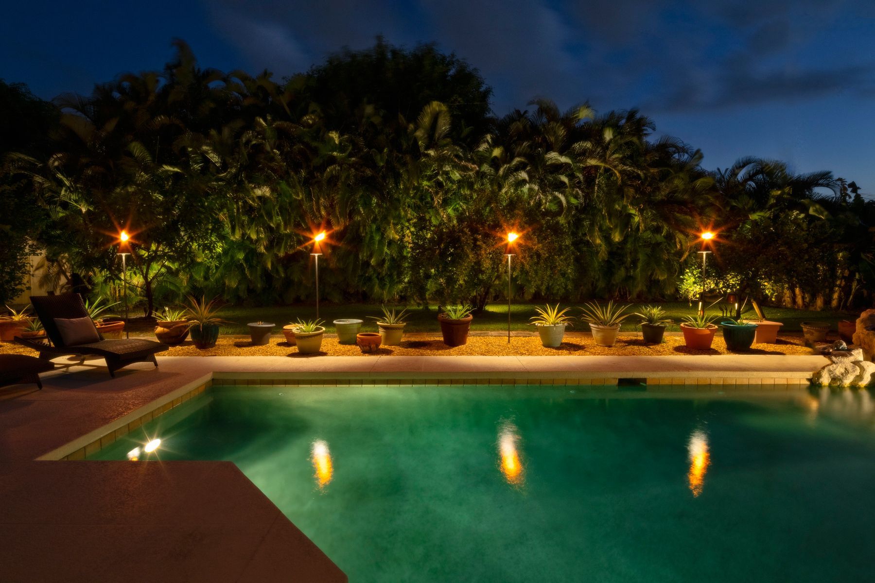 Poolside scene at dusk, lit by tiki torches and reflected in the water, with potted plants and foliage in the background.