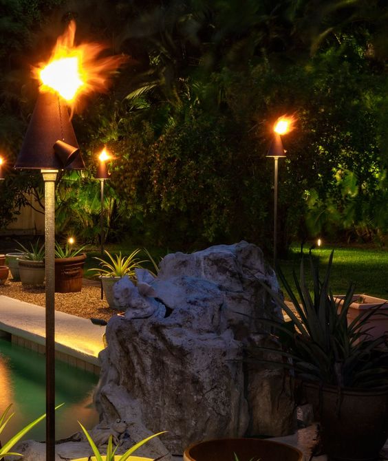 Tiki torches illuminate a nighttime backyard with plants, a rock, and a pool.