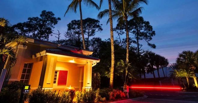 Luminous entryway to a residential area at dusk. Palm trees, warm light, and red security gate.