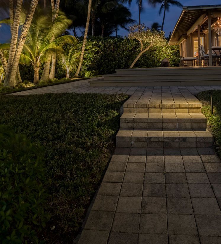 Brick pathway and steps illuminated by outdoor lighting leading to a patio with palm trees and a building in the background at dusk.