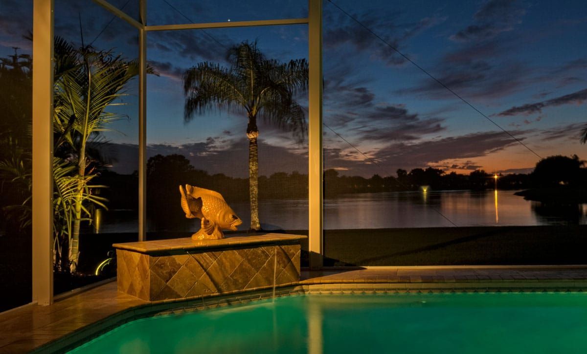 Swimming pool and pond at dusk; bronze statue on ledge, palm trees, and illuminated background.