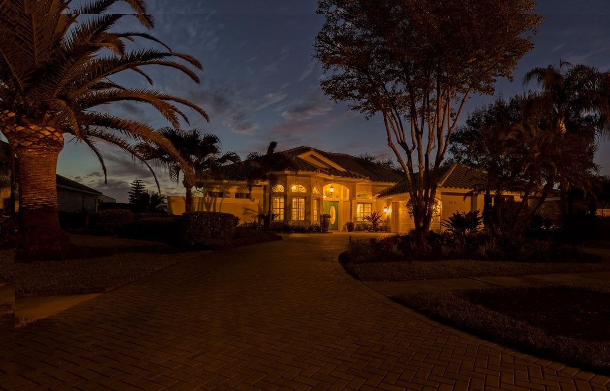 A house at dusk with lit windows, a long driveway, and palm trees.