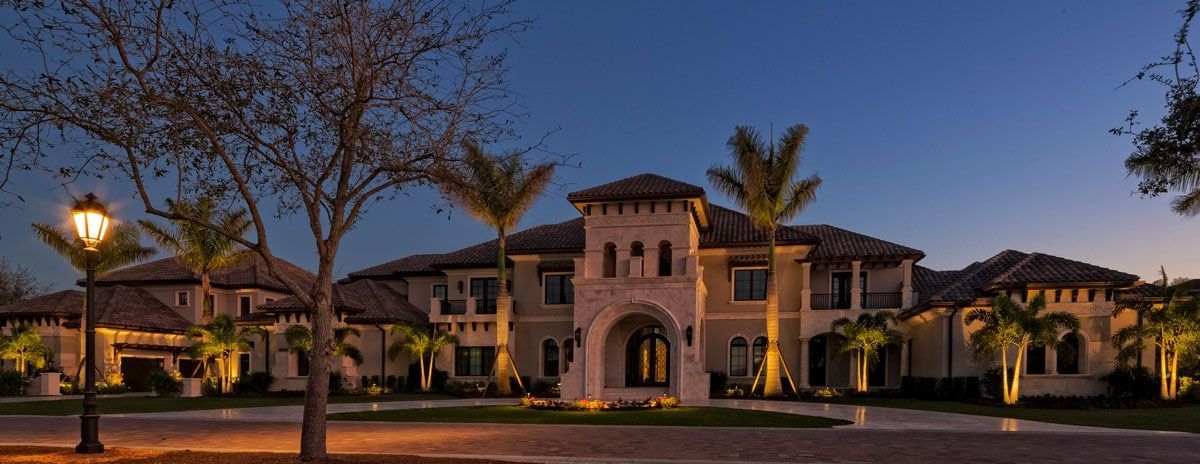 Night view of a large, lit mansion with palm trees and a lamp post.
