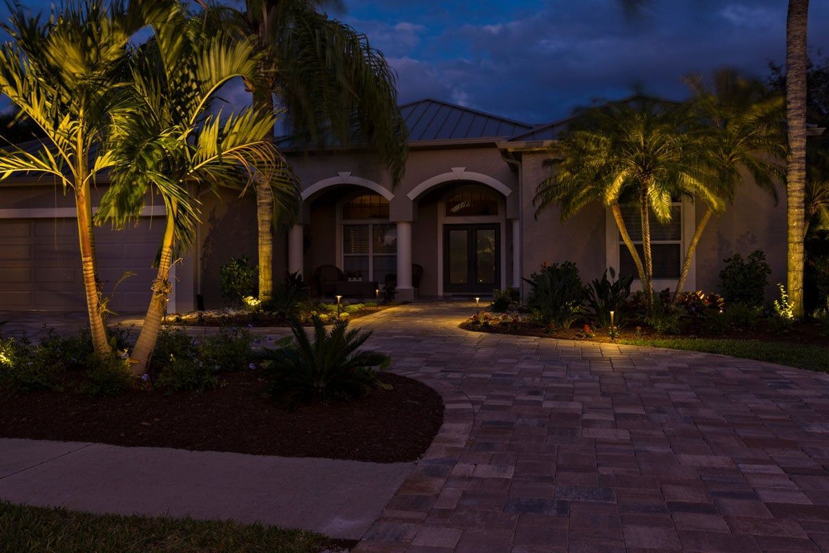 House exterior at night with palm trees illuminated by landscape lighting on a brick paver driveway.
